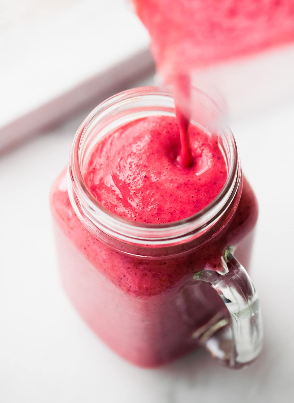 A bright pink smoothie being poured into a clear glass with handle.