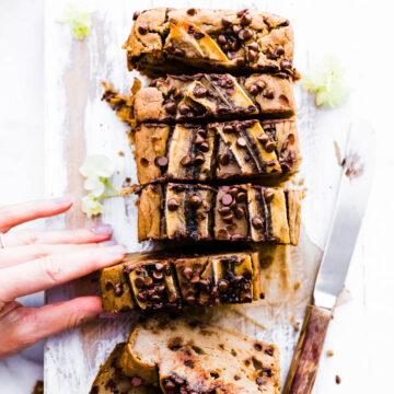 Thick slices of banana bread with chocolate chips on cutting board, a woman's hand removing one slice