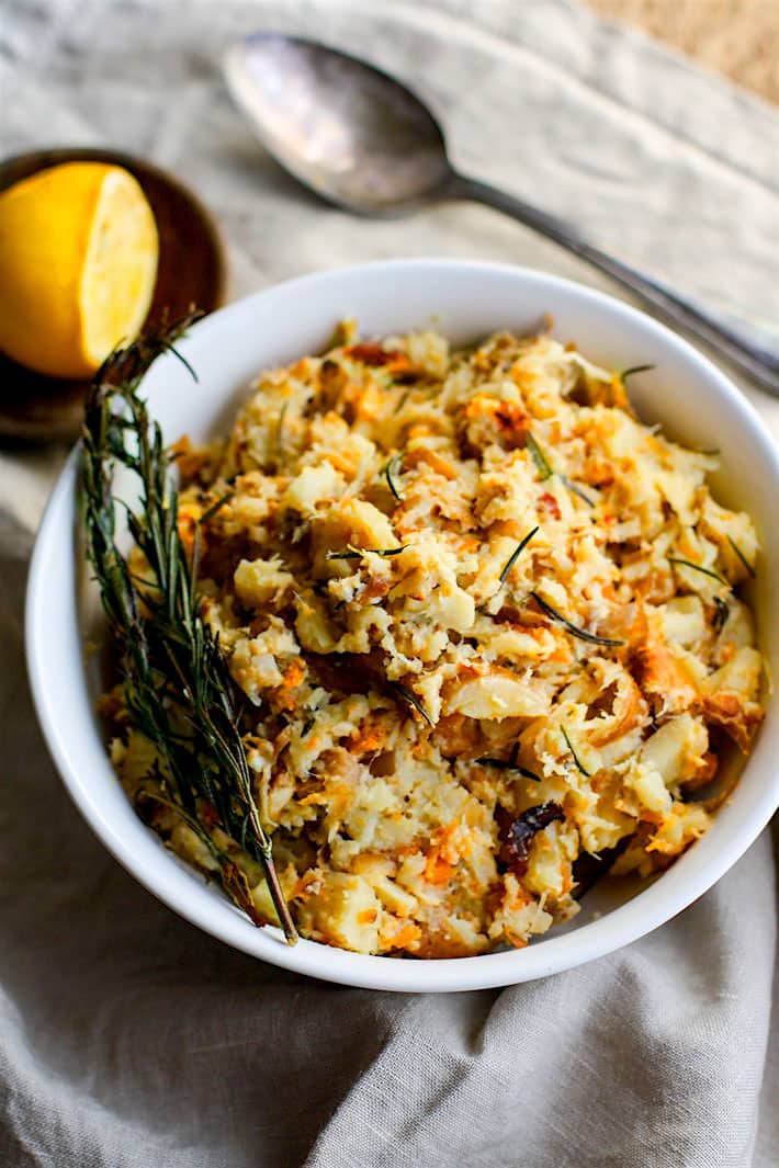 A bowl of rosemary carrot & parsnip mash sitting on a linen tablecloth with a spoon next to the bowl.