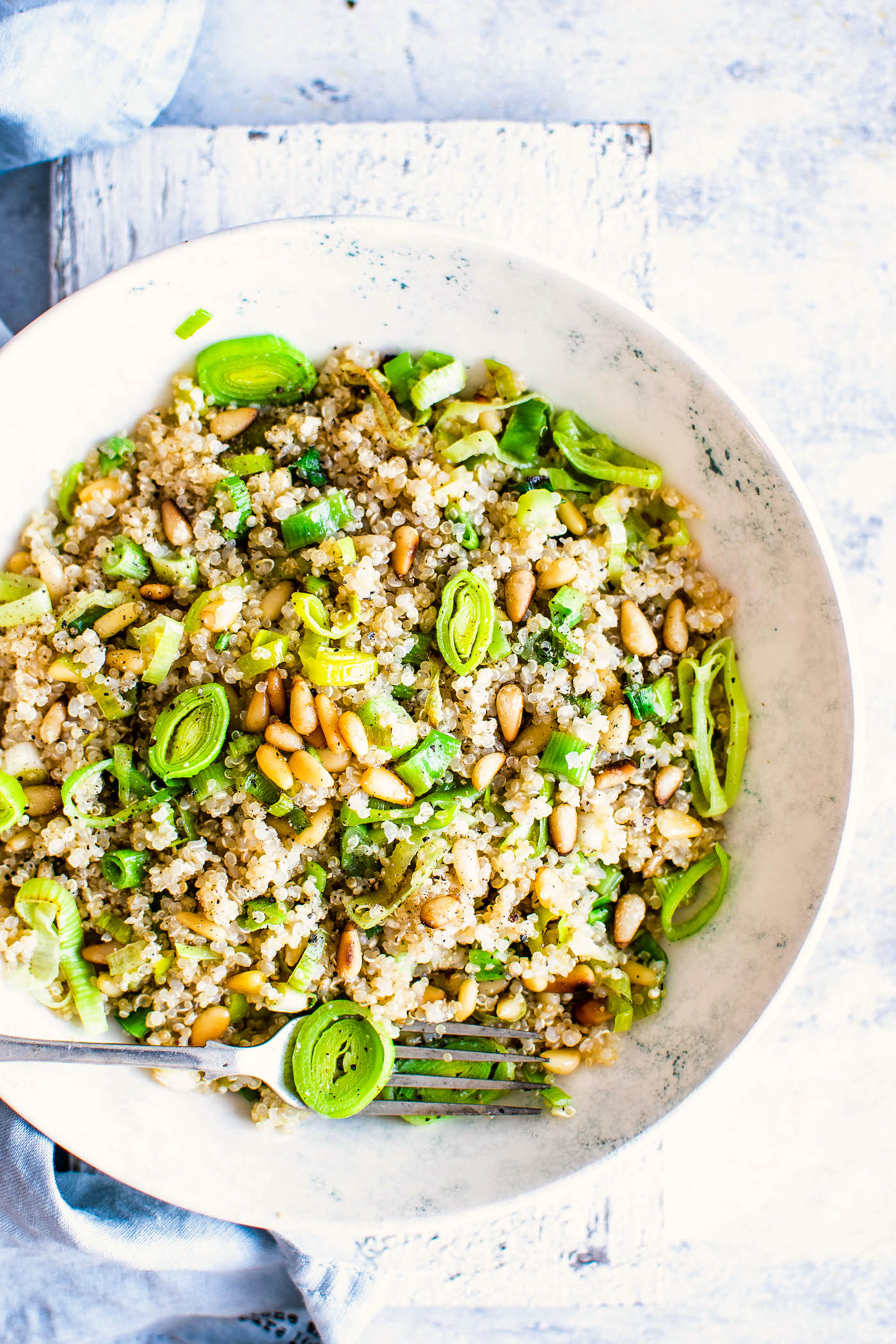 Overhead view white bowl filled with quinoa salad with pine nuts and leeks on a white cutting board.