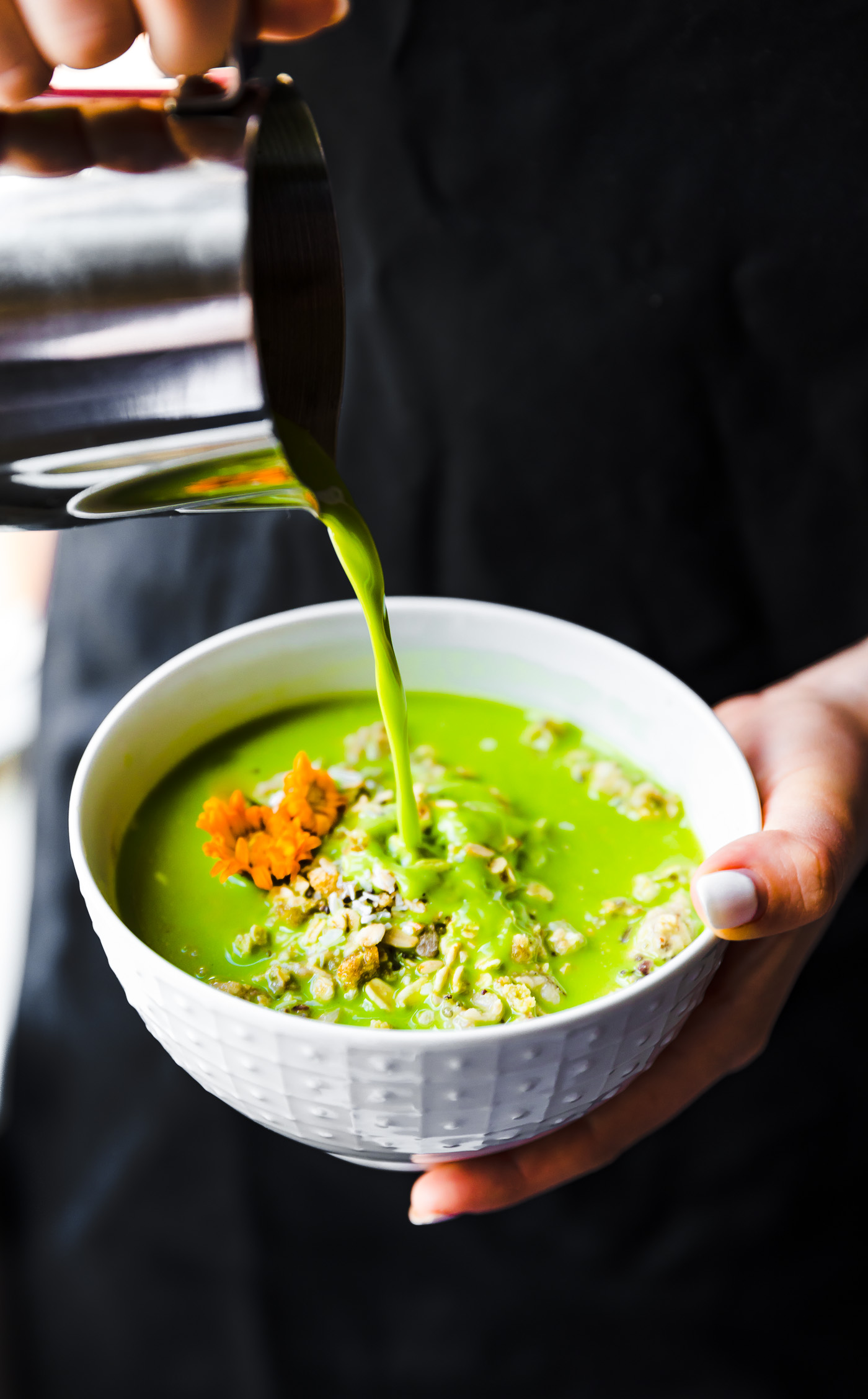 A white bowl filled with green tea matcha oatmeal being held in one hand, a silver pitcher pouring in matcha milk
