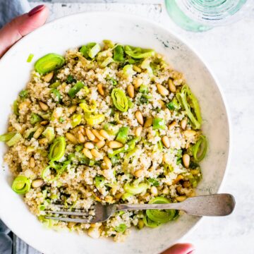 Two hands holding a white bowl filled with toasted pine nut quinoa salad with leeks.