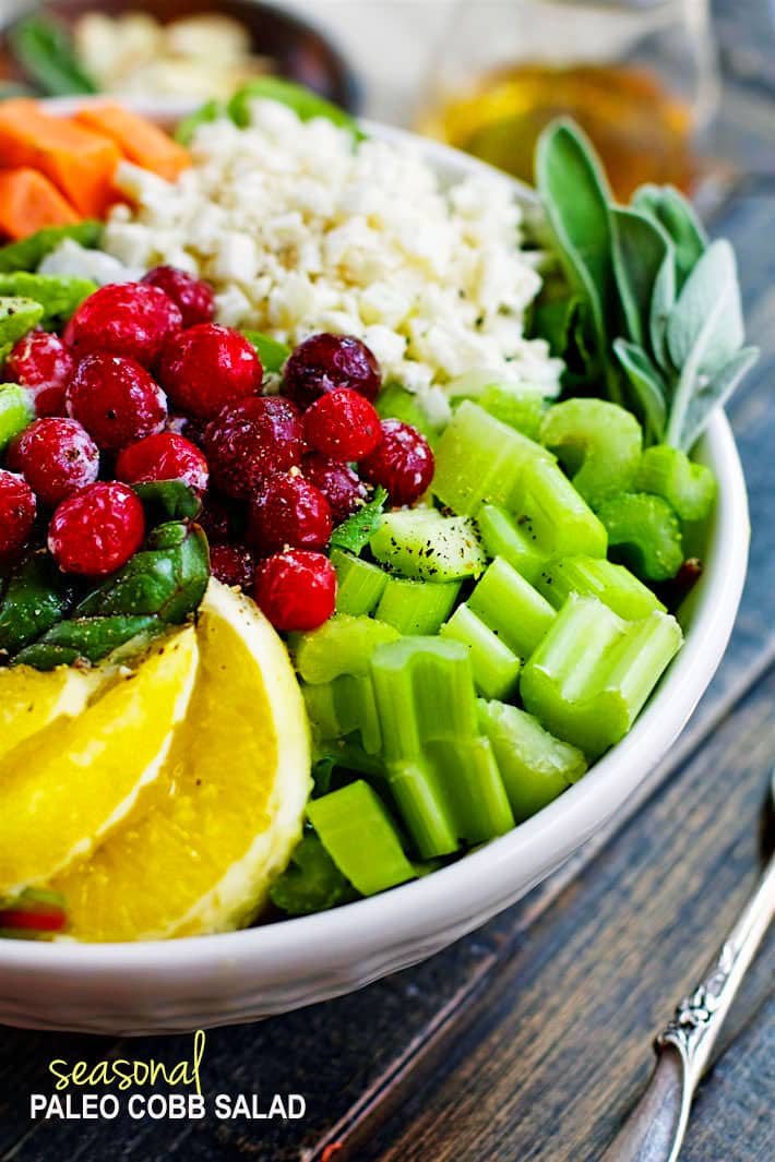 Seasonal Cobb Salad in a white bowl with piles of celery, cranberries, cauliflower rice, sage, and fresh lemons ready to be mixed together.