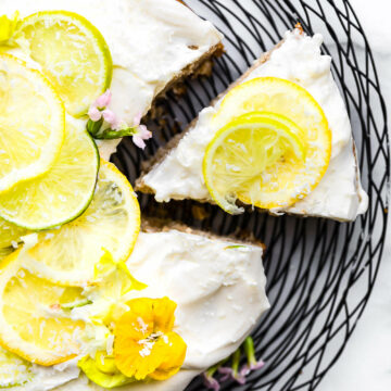 Overhead view citrus coconut vegan cake with white frosting and thin slices lemon and lime on black wire rack, one slice pulled away from cake.