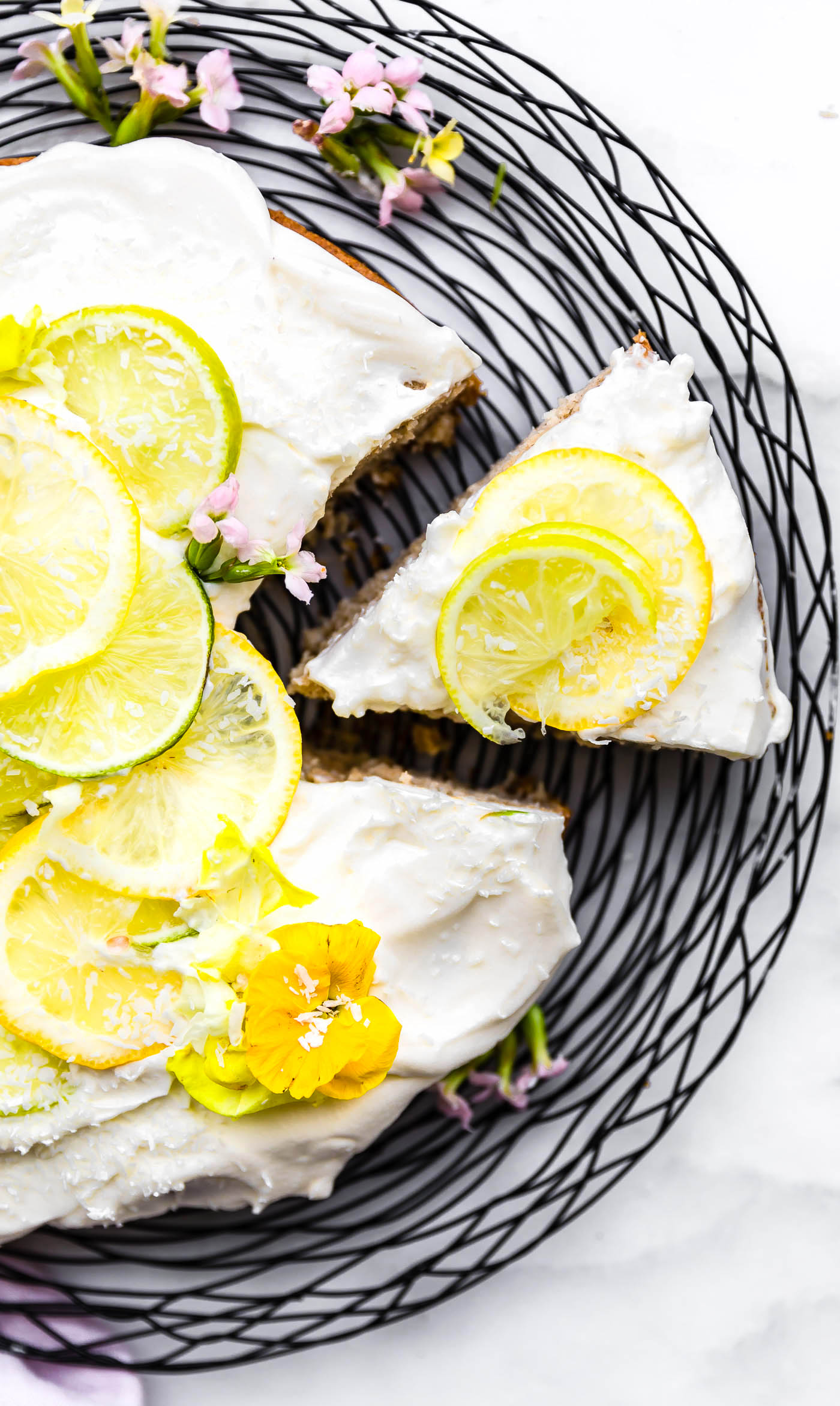 Overhead view citrus coconut vegan cake topped with coconut cream frosting with lemon slices, one slice pulled away from cake.