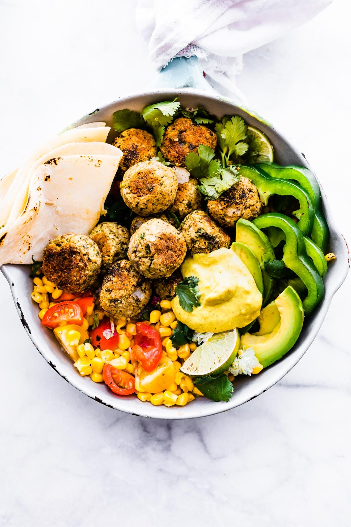 Overhead view white metal serving bowl filled with Mexican falafel balls, fresh cut vegetables and tortilla wrap folded.