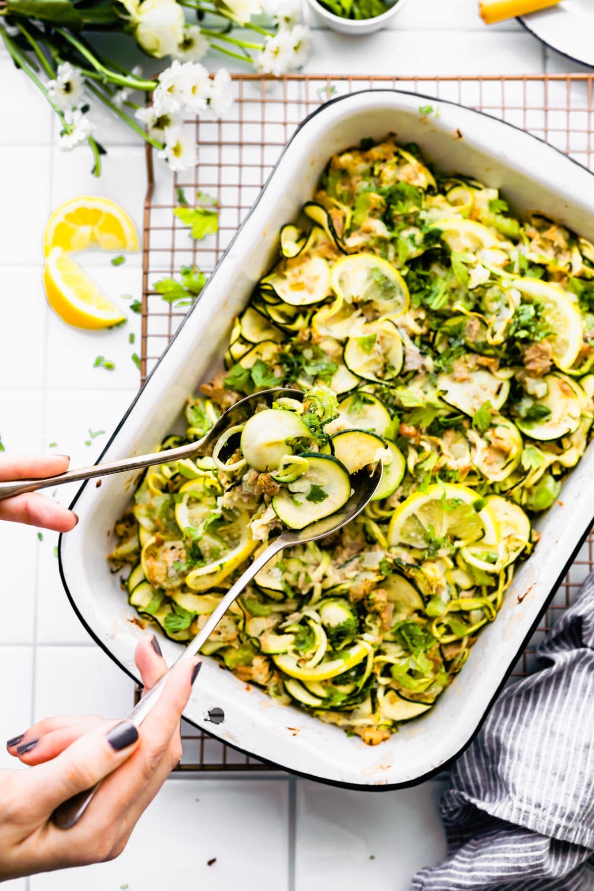Paleo Tuna Green Chile Zoodle Casserole on white board with plate above and flowers, lemon, herbs on side