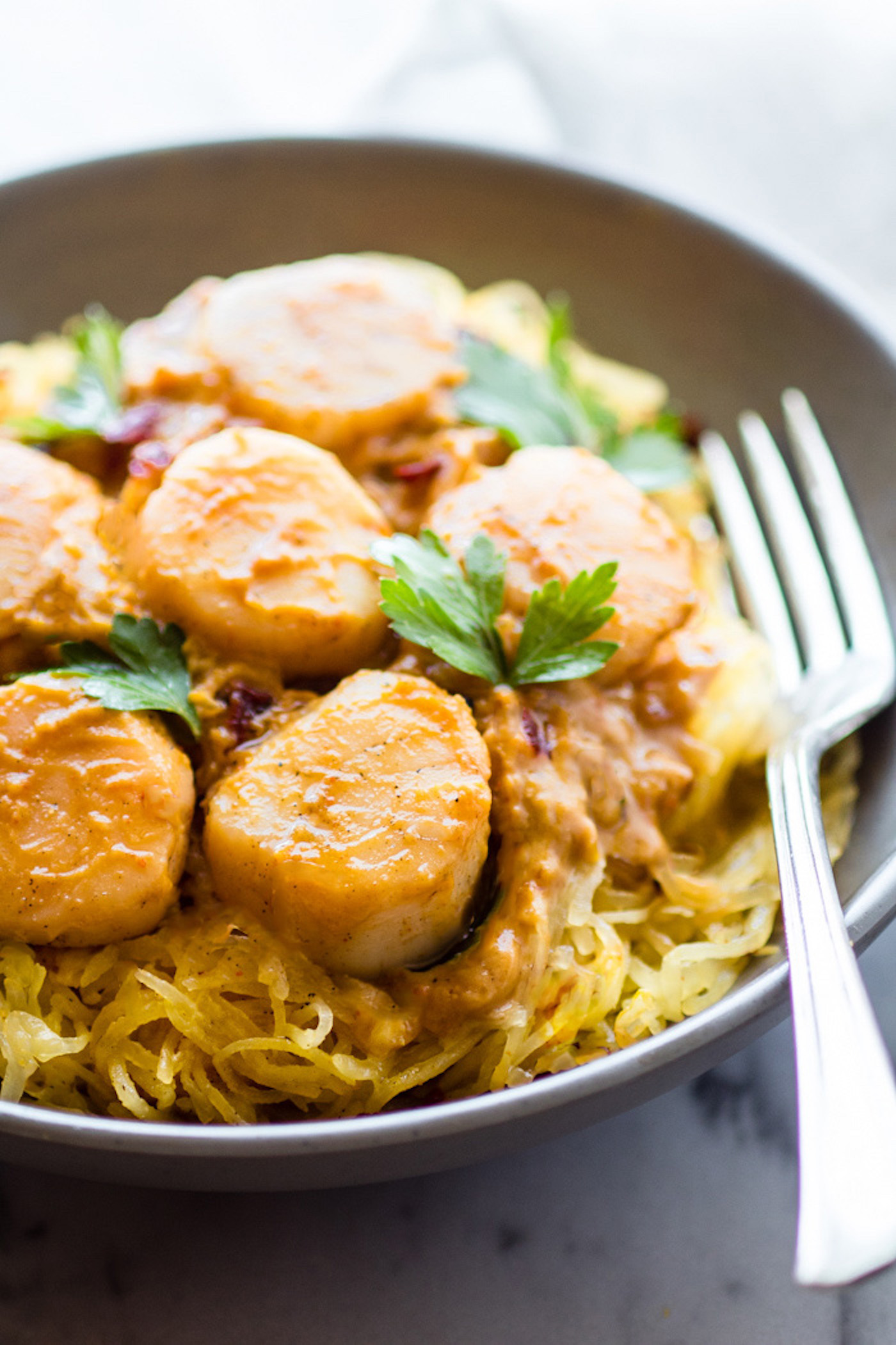 Spaghetti Squash Pasta makes for an easy paleo pasta alternative! This gluten-free Seafood Pumpkin Spaghetti Squash Pasta is topped with a miso pumpkin sauce and buttery scallops. A healthy lower carb version of your favorite pasta dish. DIG IN! #spaghettisquashpasta #lowcarbrecipes #seafoodrecipes Close up view of miso pumpkin sauce coated scallops over spaghetti squash with fresh herbs.