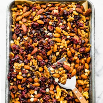 Pumpkin spice trail mix spread on a parchment-lined sheet pan with almonds, walnuts, dried cranberries, and chocolate chips, with a spatula scooping a portion.