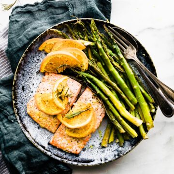 salmon on blue plate with orange slices on top and herbs. blue linen on the side. Fork on plate to the right of salmon. overhead angles shot