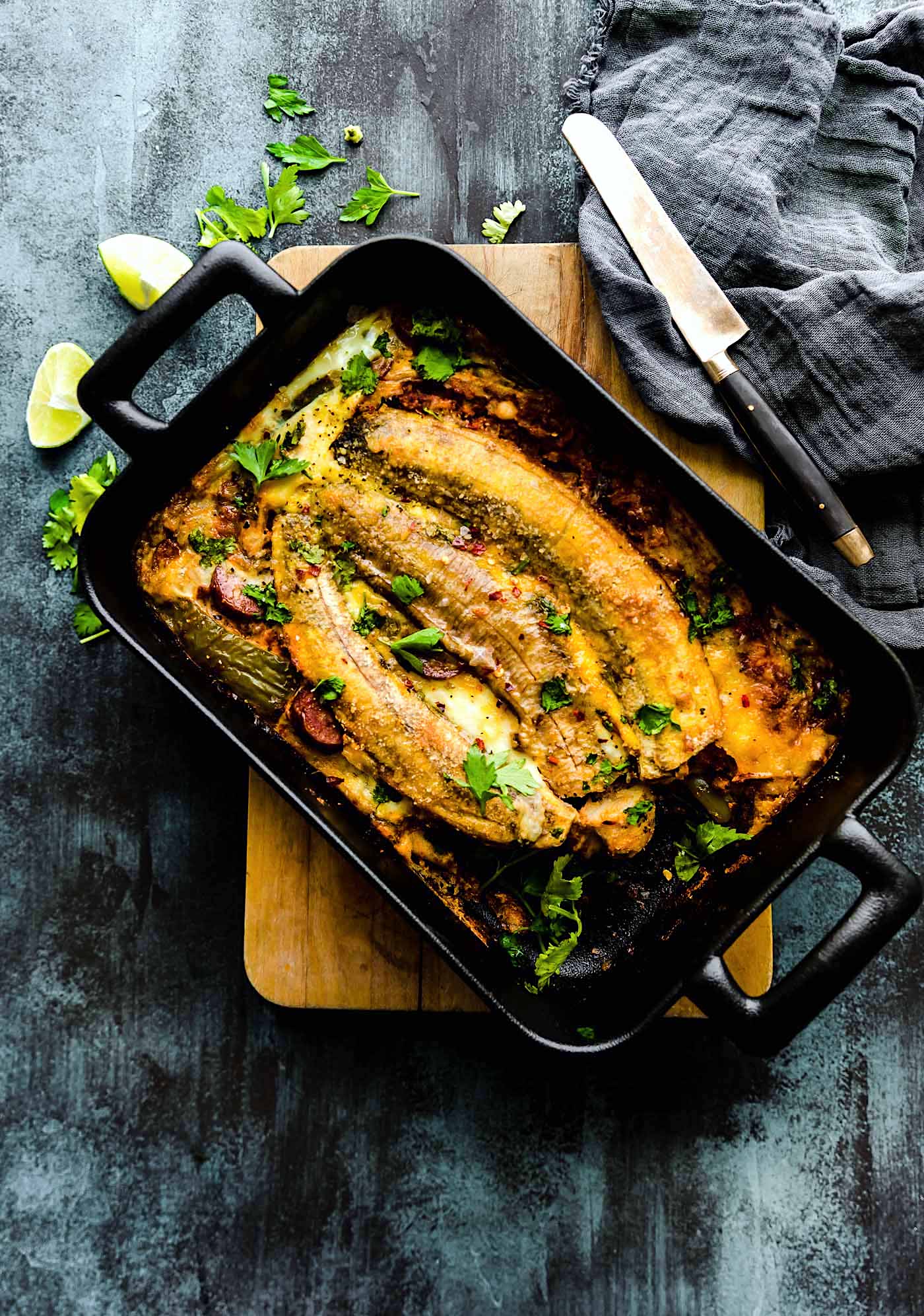 Breakfast lasagna topped with plantains in black loaf pan on wooden cutting board.