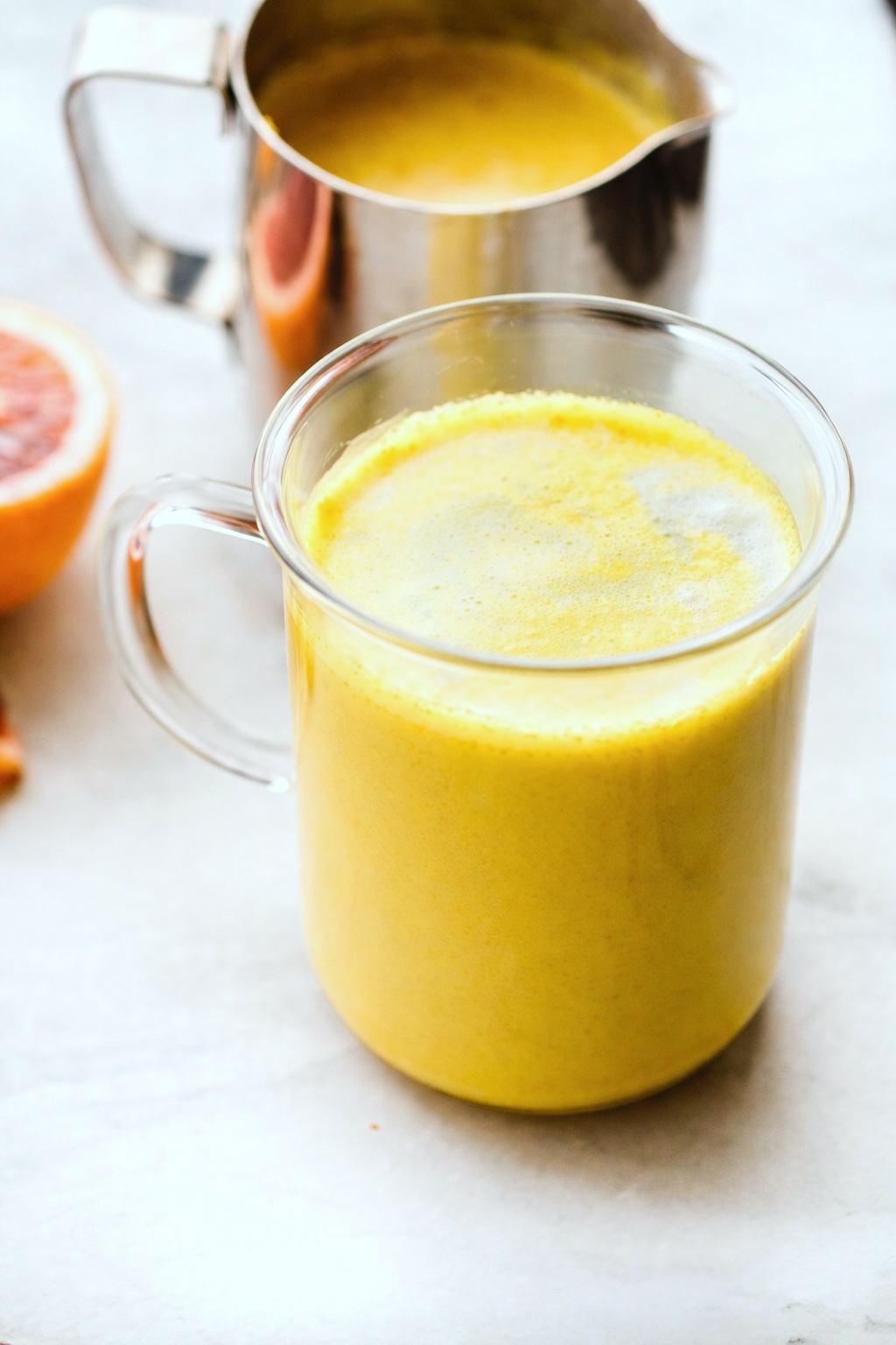This Orange Turmeric Tea Latte will boost your health and energy! It's a cold-busting vegan latte, loaded with extra Vitamin C, brain-boosting healthy fats, minerals, and anti-inflammatory properties. Caffeine-free. #turmerictea #veganlatte #antiinflammatory #recipes Golden turmeric latte in a glass mug with metal pitch of extra latte behind it