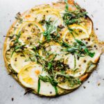 Overhead view socca pizza topped with lemon slices and fresh herbs on silver baking sheet.