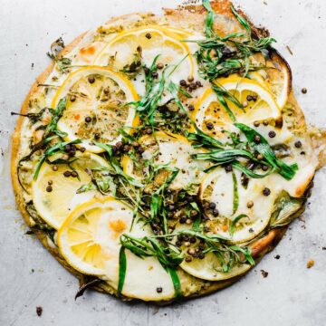 Overhead view socca pizza topped with lemon slices and fresh herbs on silver baking sheet.