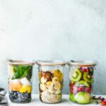 Side view three glass jars filled with fresh fruit, vegetables, grains, and seeds against turquoise background.
