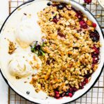 Coconut Summer Berry Crisp in metal pie plate on cooling rack, melting ice cream on half of crisp