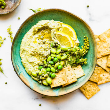 Overhead view vegan edamame pesto dip in green bowl with crackers, edamame, and lemon slices.