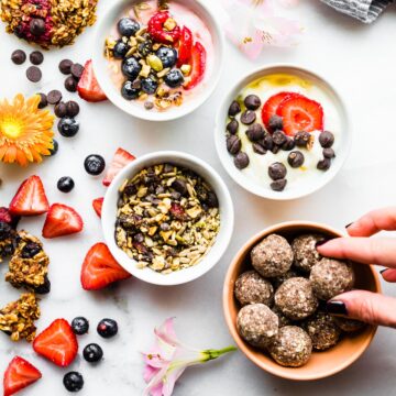Three bowls filled with yogurt topped with trail mix, energy bites in bowl, and breakfast cookies stacked up.