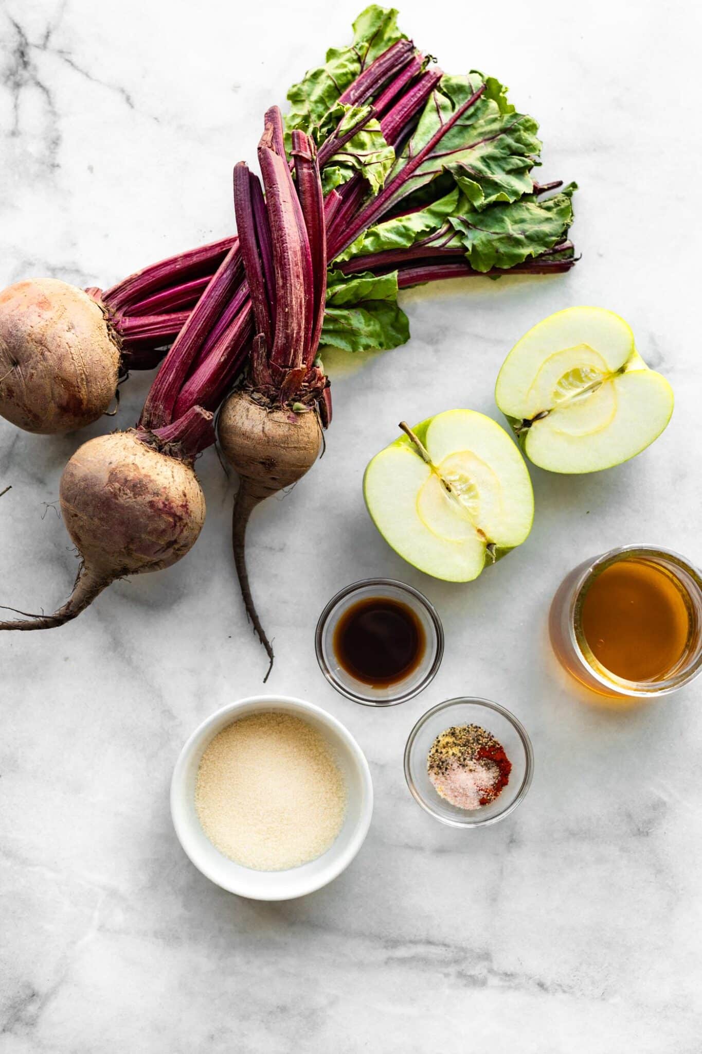 whole beets, sliced green apple, spices, and oil in bowls on a white marble background