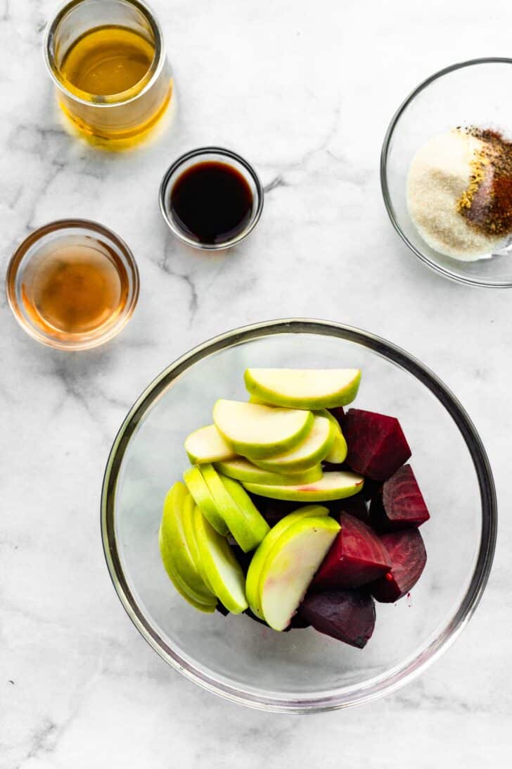 chopped apple and beets in a clear bowl with oil and spices in other bowls around it