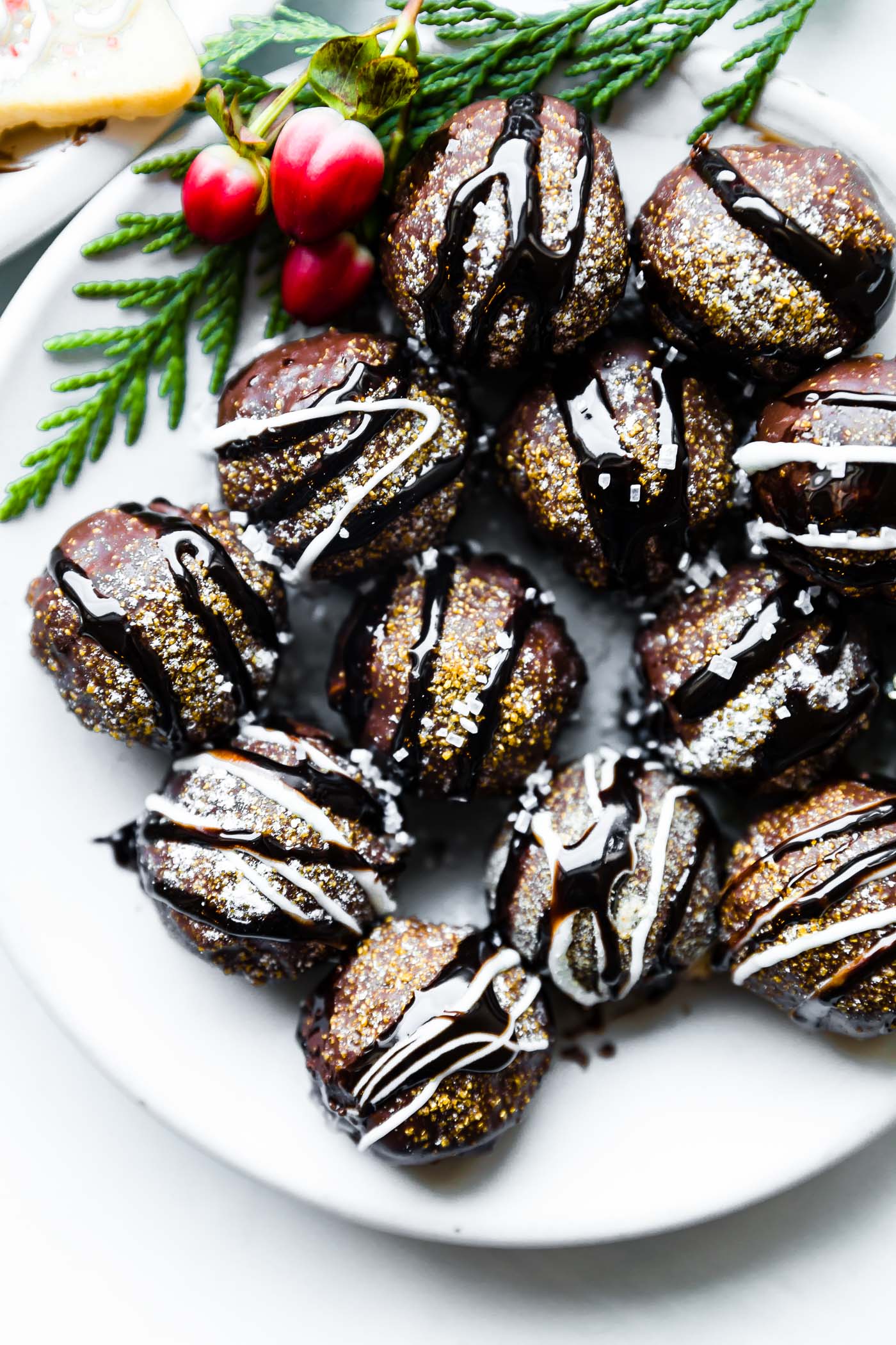 Several no bake chocolate covered cookie truffles with dark chocolate drizzled over top on a white platter with red berries and evergreen on the side.