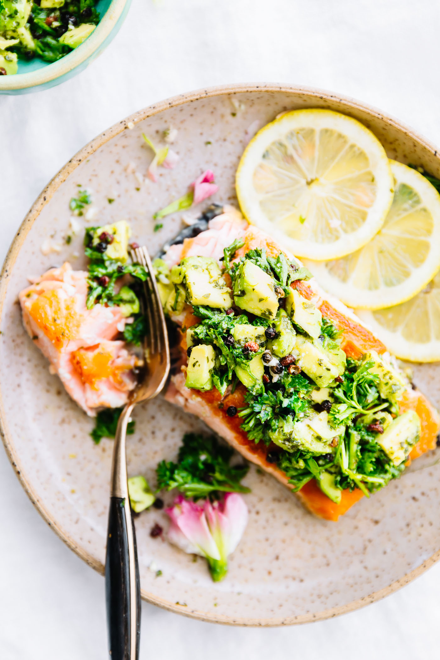 Salmon fillet on brown speckled plate, topped with avocado gremolata and lemon slices.
