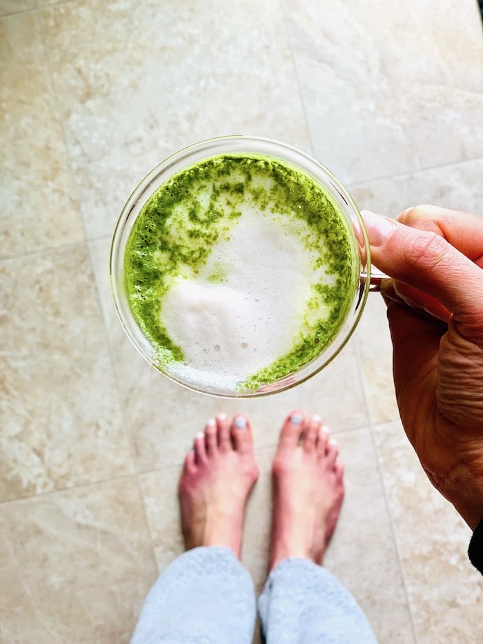 A woman standing and holding a matcha latte over the floor with her feet showing.