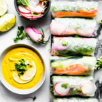 Several spring rolls lined up on black cutting board, ginger curry dipping sauce on the side.