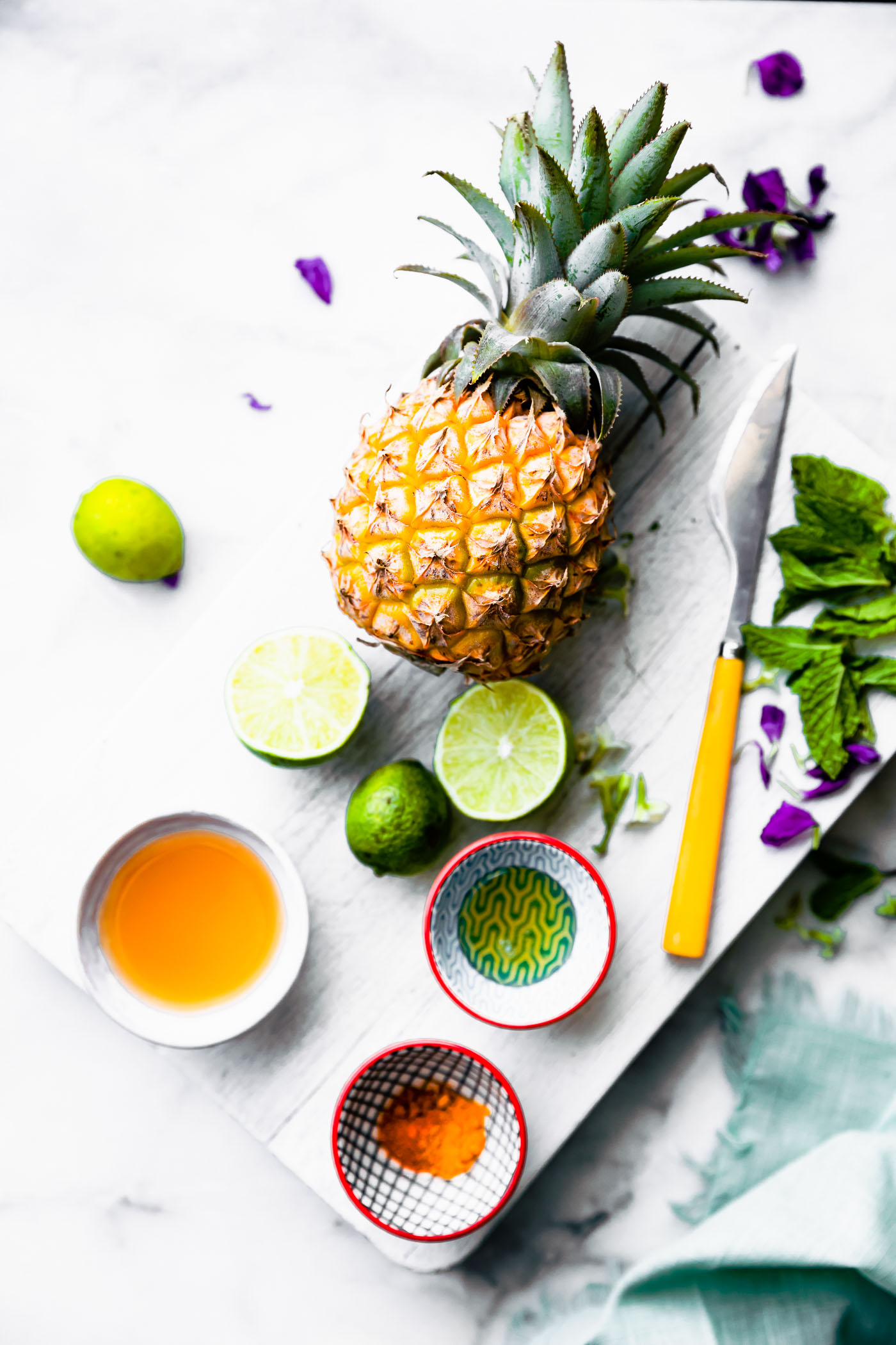 A fresh pineapple, lime halves, knife, and apple cider vinegar on a white cutting board.