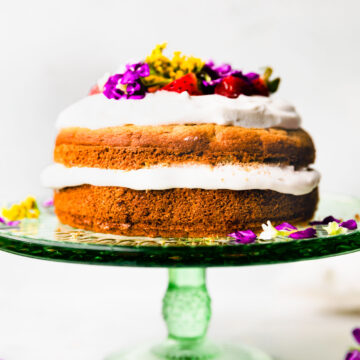 Two layered birthday for dog with white yogurt frosting, topped with fresh flowers and strawberries, on green cake stand.