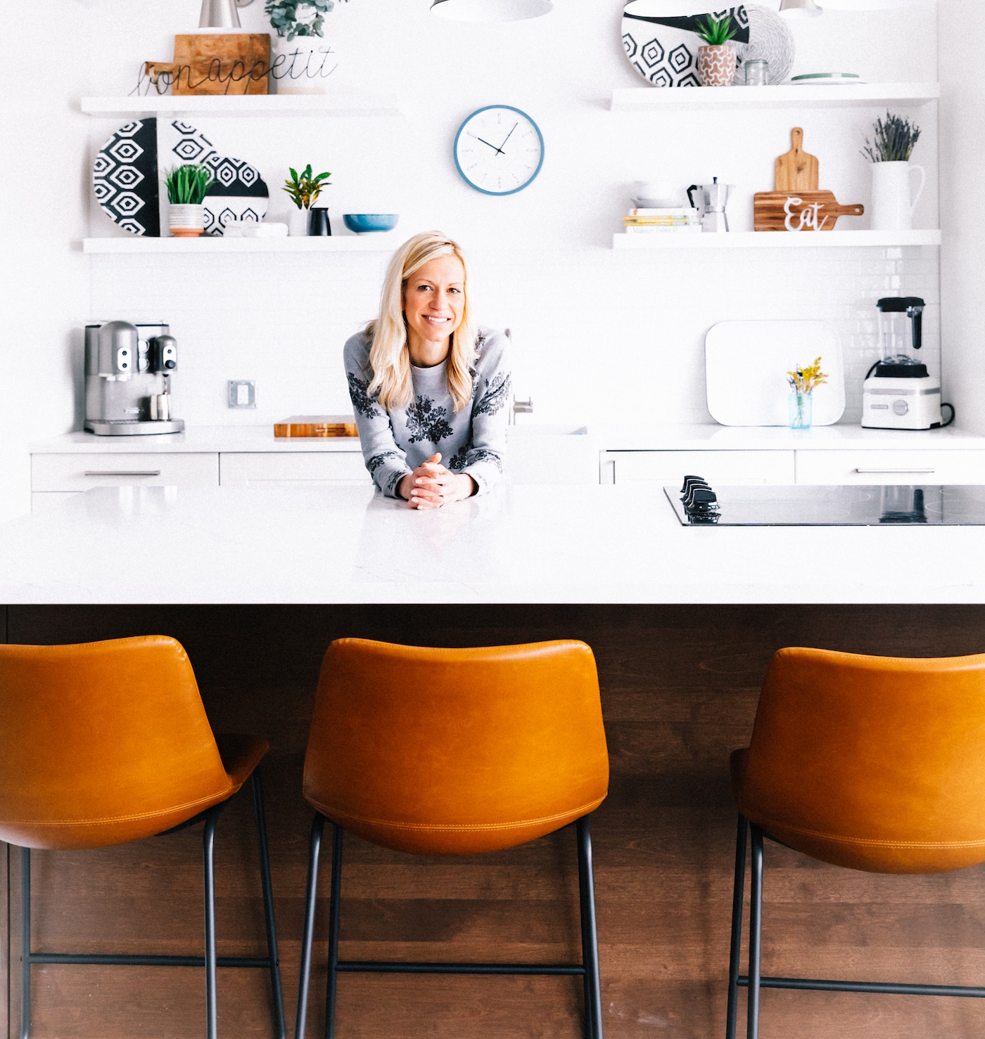A woman standing behind white kitchen island resting forearms on counter, white countertops and floating shelves behind.