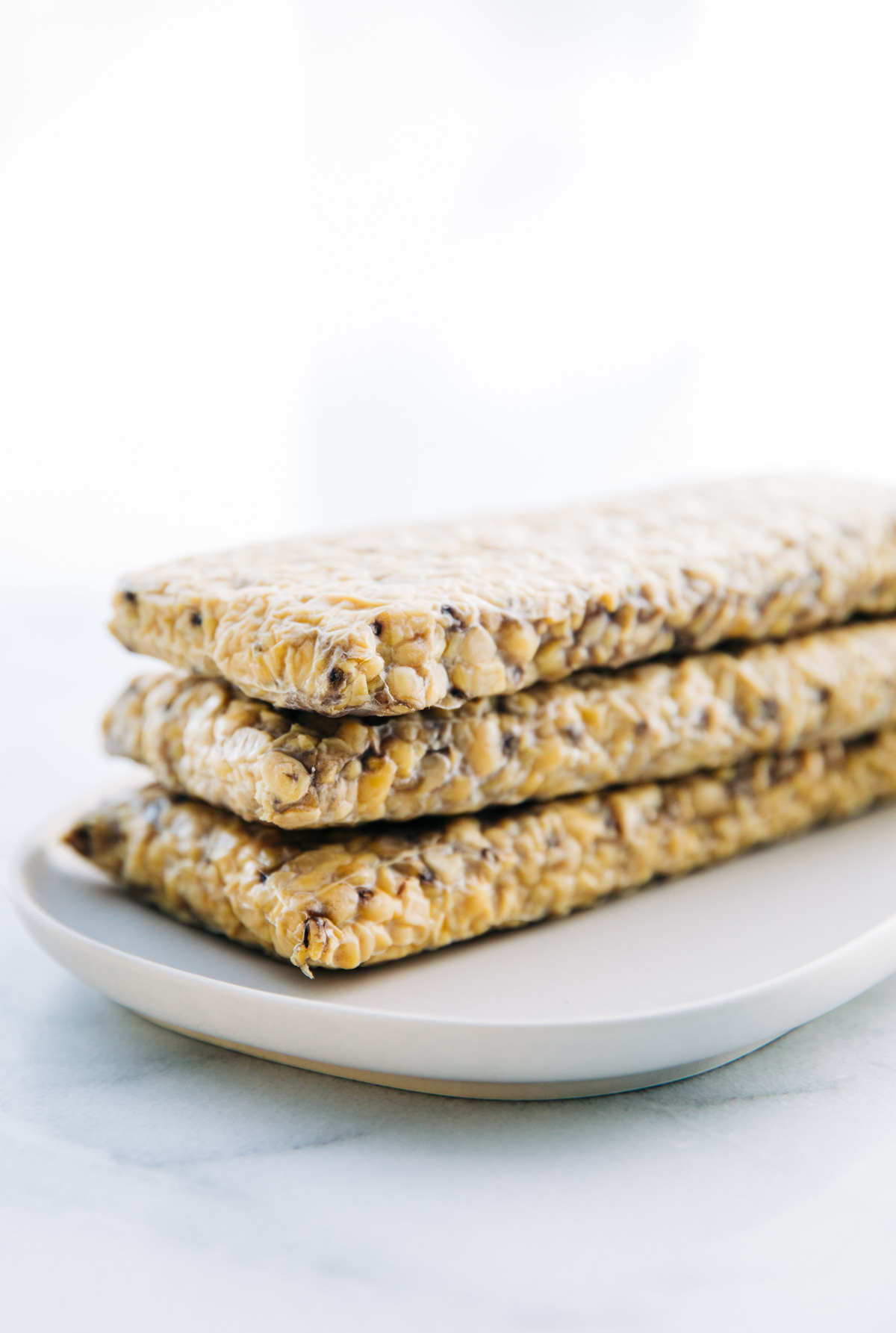 Three raw tempeh slabs stacked on top of each other on a plate.