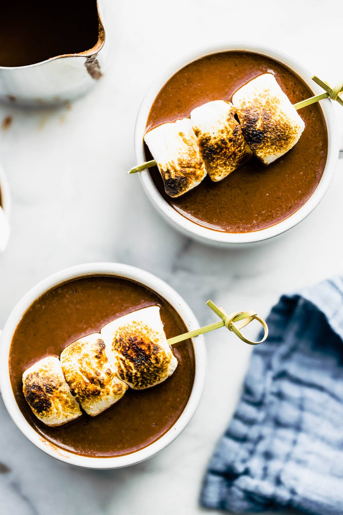 Overhead photo of two boozy hot chocolate drinks with skewers of toasted marshmallows on top.
