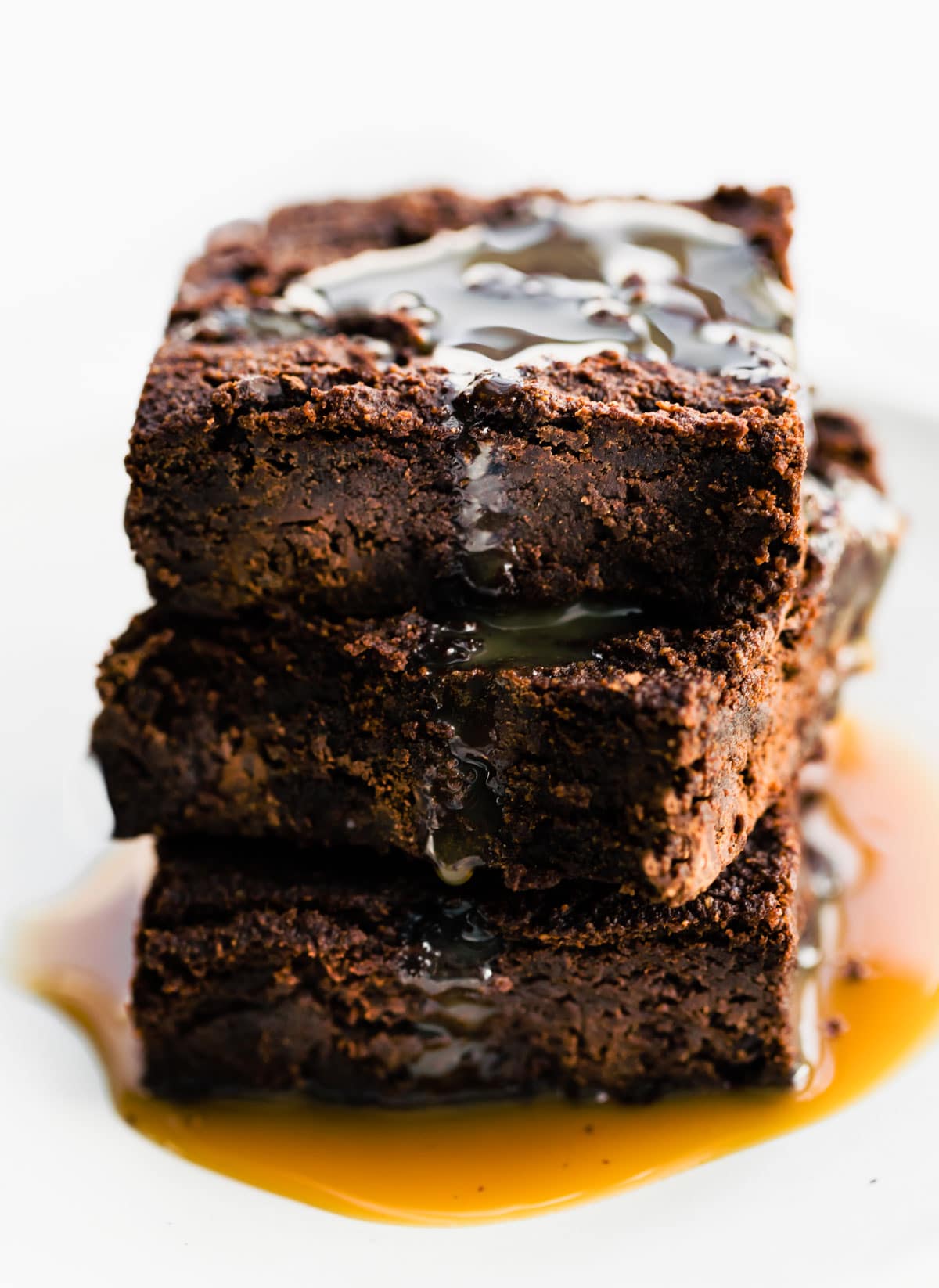 Three vegan brownies with salted caramel sauce drizzled on top stacked up on each other on a white plate.