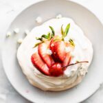 An overhead image of a pavlova on a white plate, topped with fresh strawberries and a cherry.