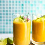 A side shot of two turmeric smoothies in glasses on a serving tray with a garnish of lime, mint, and flowers.