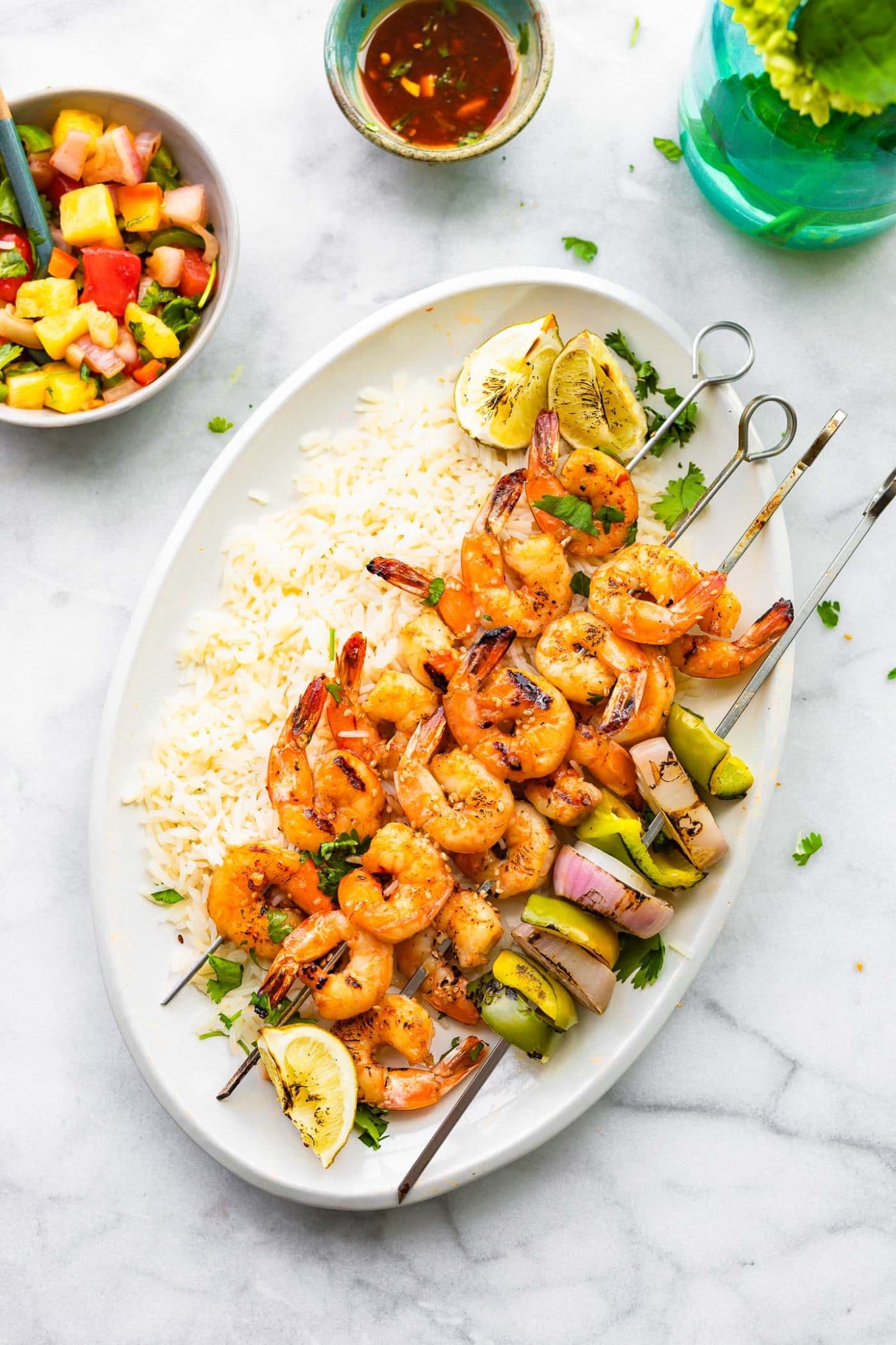 Overhead image of grilled skewered shrimp on a platter of rice with a side of watermelon salsa.