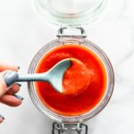 Overhead image of a spoonful of homemade sriracha sauce being lifted out of a storage jar.