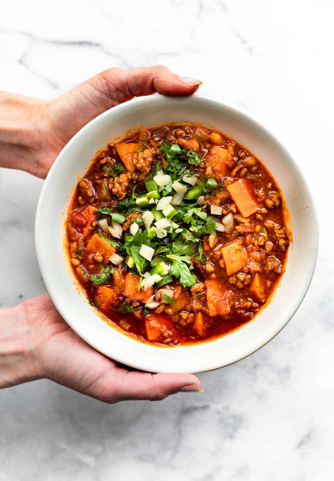 two hands holding a white bowl of no-bean sweet potato turkey chili with mole sauce topped with onions and cilantro