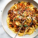 a white bowl full of long pasta noodles topped with vegan mushroom bolognese and grated cheese with a spoon and a fork gathering a bite to eat