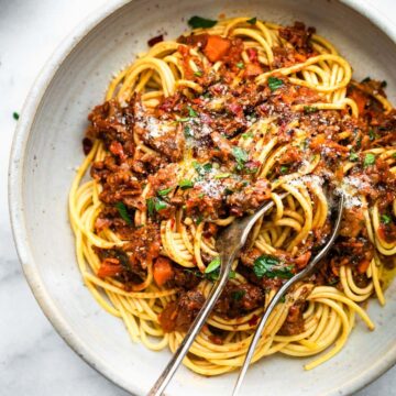 a white bowl full of long pasta noodles topped with vegan mushroom bolognese and grated cheese with a spoon and a fork gathering a bite to eat
