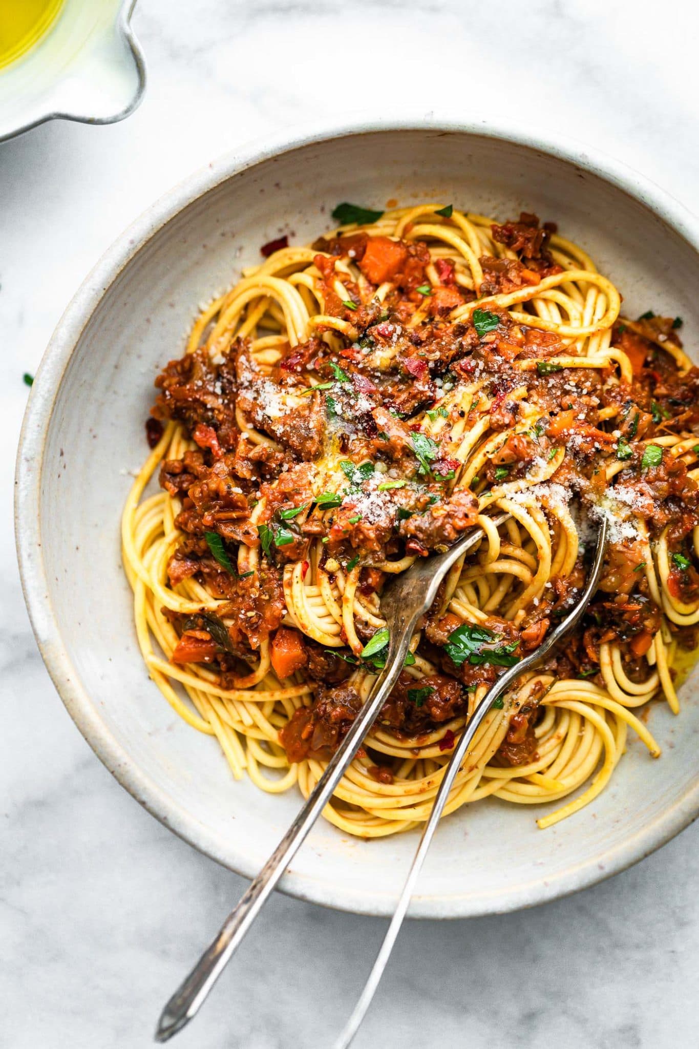 a white bowl full of long pasta noodles topped with vegan mushroom bolognese and grated cheese with a spoon and a fork gathering a bite to eat