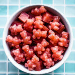 a white bowl on a counter with a blue tiled background filled with red colored homemade gummies with probiotics in the shape of cubes and bears