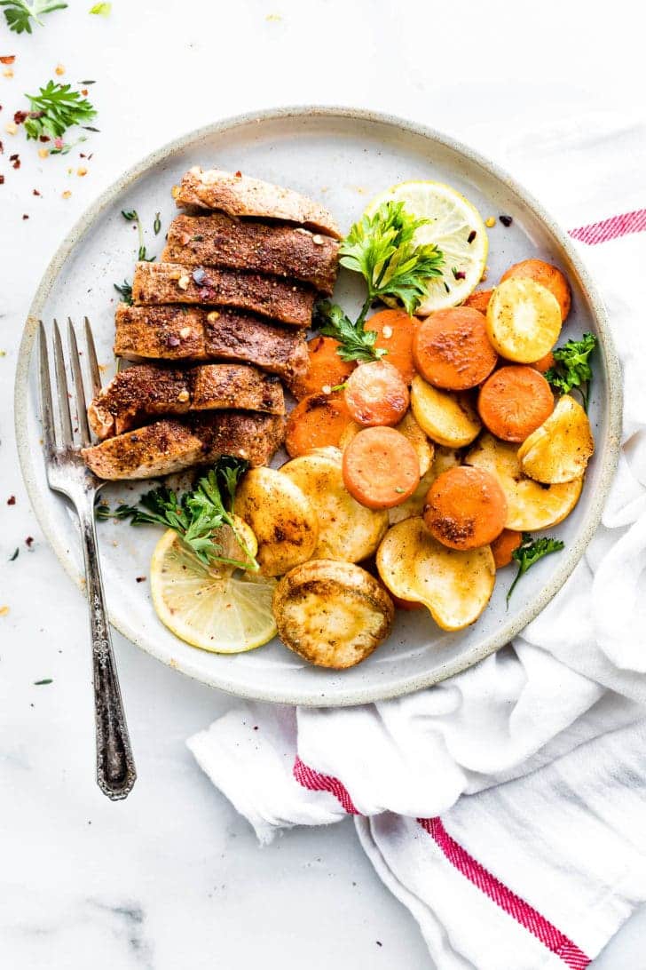 cinnamon chipotle spice rubbed pork chop sliced into pieces on a white plate with roasted carrots and parsnips on the side with a fork and lemons and herbs for garnish
