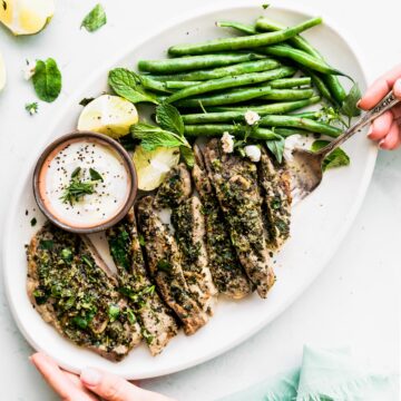 a white serving platter filled with broiled lamb chops with mint chimichurri sauce, green beans, a halved lime, and a white yogurt dipping sauce with a woman's hand holding the platter and another hand holidng a fork underneath a lamb chop