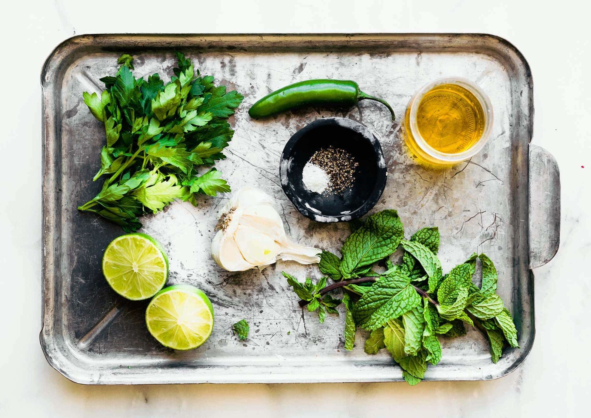 a serving tray with a cup of olive oil, fresh parsley, fresh mint, a jalapeno pepper, a black bowl with salt and pepper, garlic cloves, and a halved lime