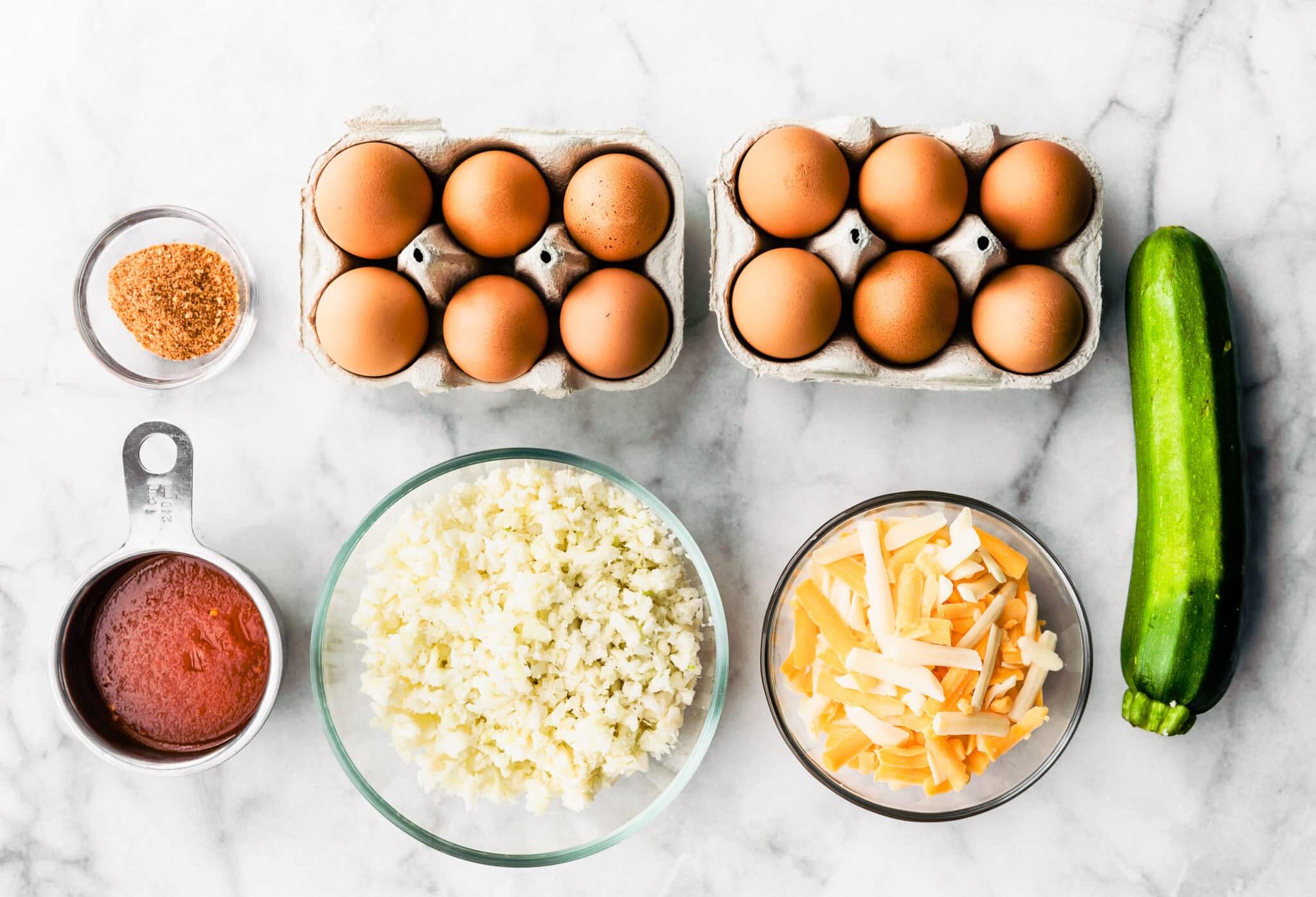 overhead image of two cartons of 6 eggs, a glass bowl with spices, a measuring cup holding enchilada sauce, a glass bowl holding riced cauliflower, a glass bowl holding shredded cheese, and a whole zucchini needed to make tex-mex egg and cauliflower casserole