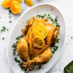 an overhead image of a whole chicken on a white serving platter topped with fresh lemon slices and surrounded by herbs