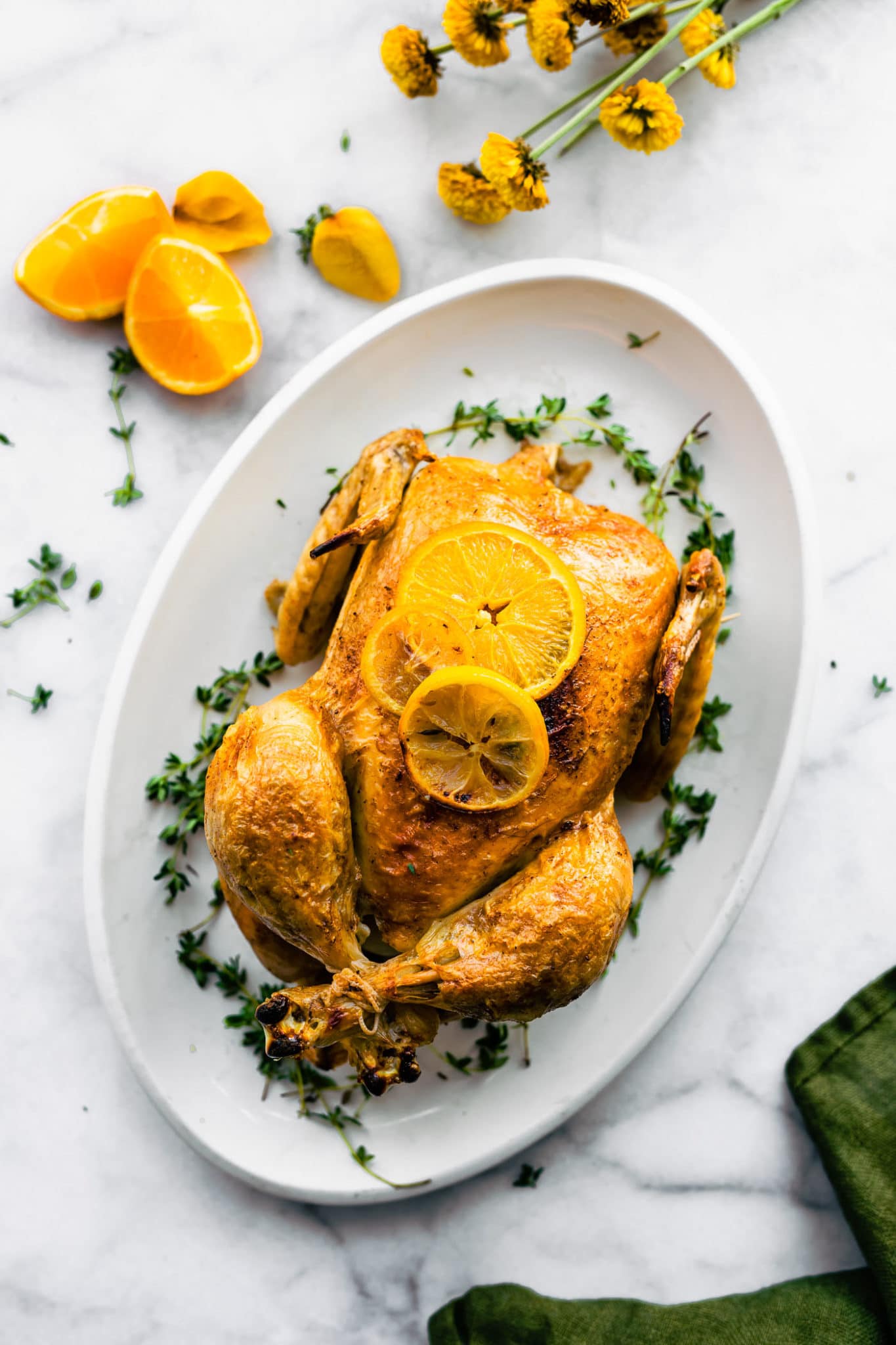 an overhead image of a whole chicken on a white serving platter topped with fresh lemon slices and surrounded by herbs