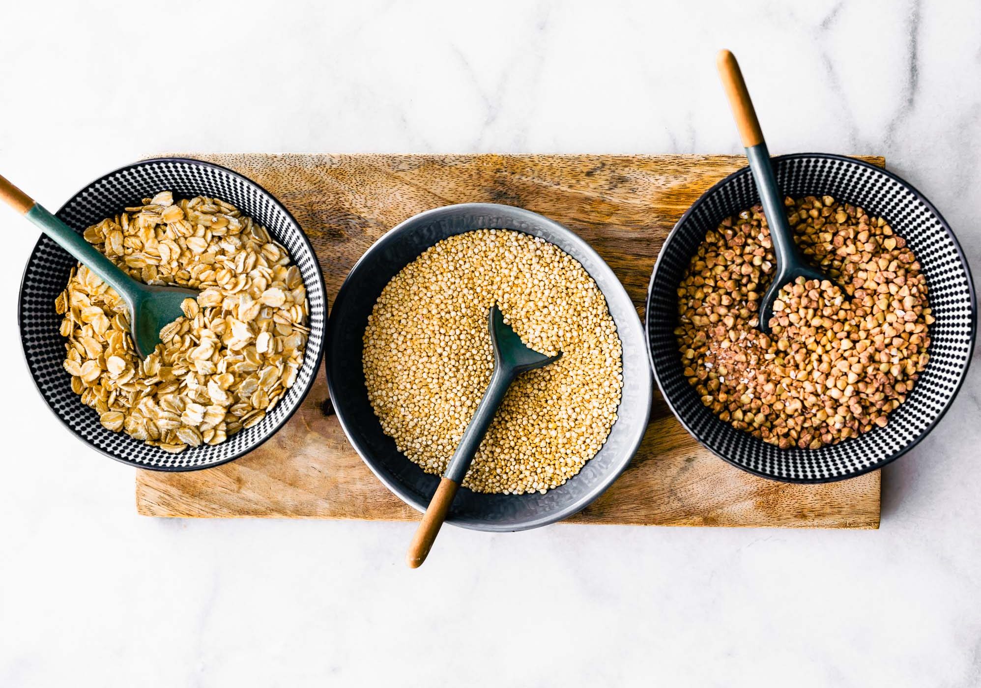 overhead image of three bowls on a wooden plank one filled with gluten free oats, one filled with millet, and one filled with buckwheat